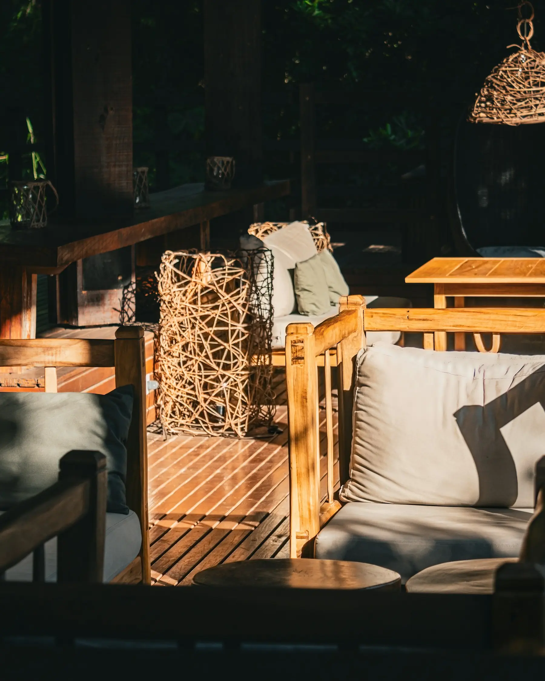 A shaded timber deck with cushioned chairs and a woven lamp overhead.