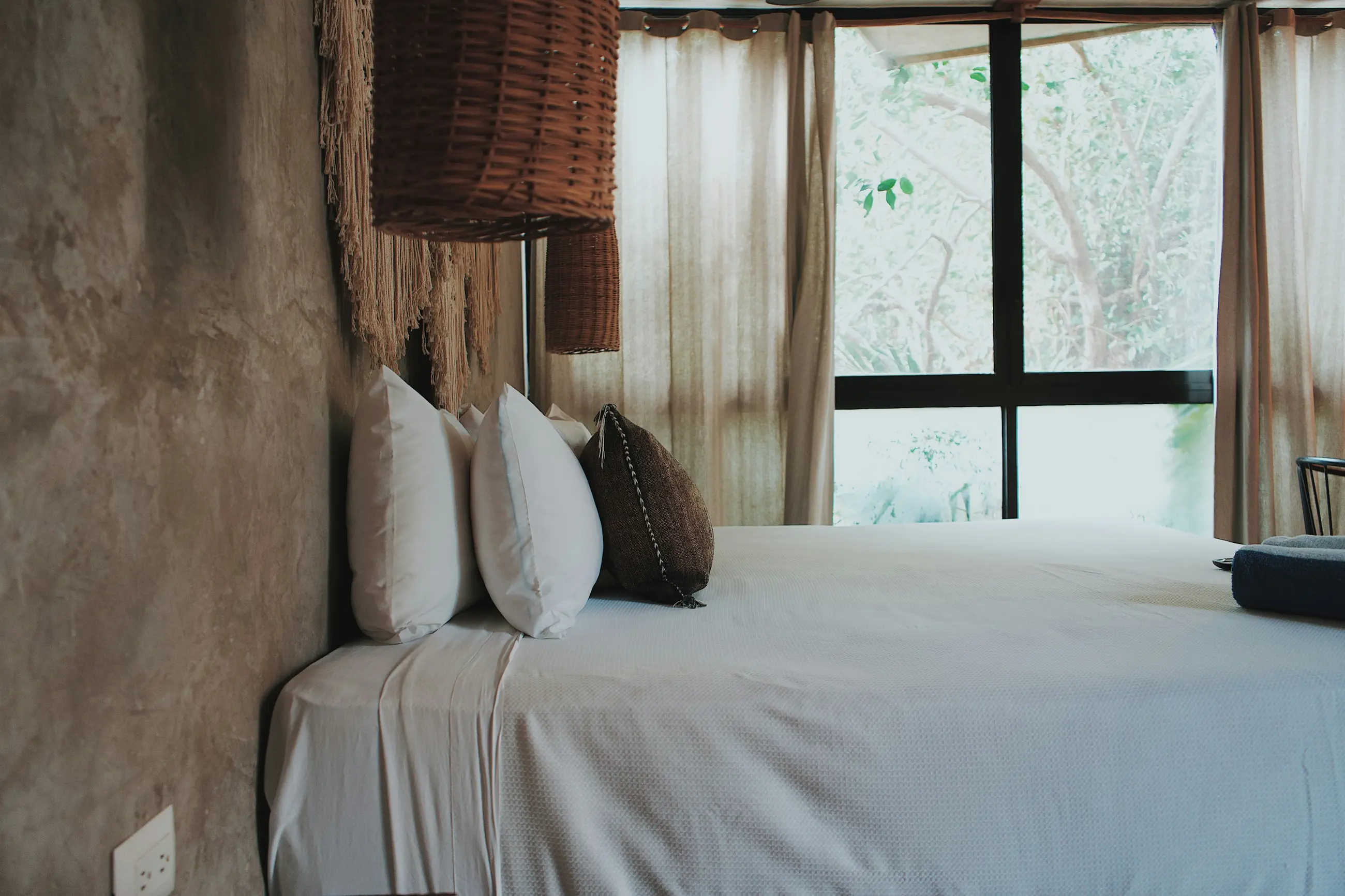 A bed made up in white beside a tall window with green pressing against it.