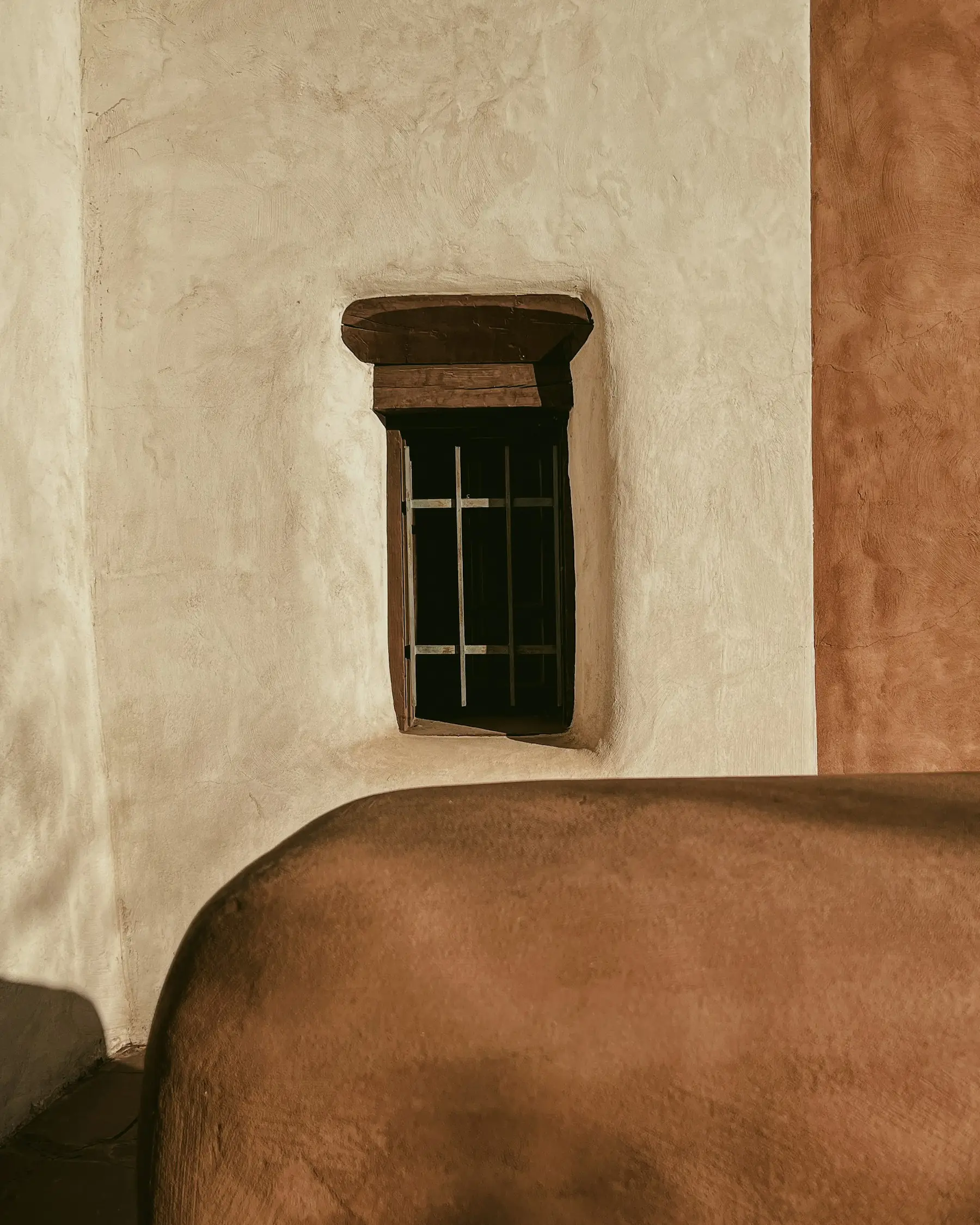 A small timber window set into a thick plaster wall, beside the corner of a bed.