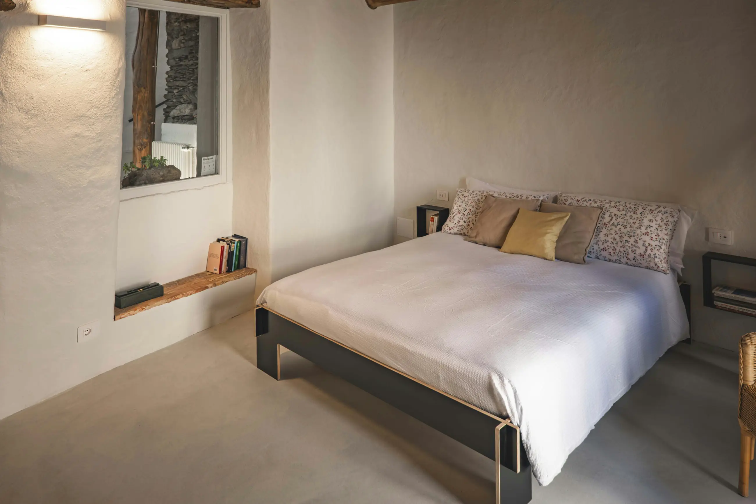 A plain bed in a whitewashed room, books stacked in a deep window niche.
