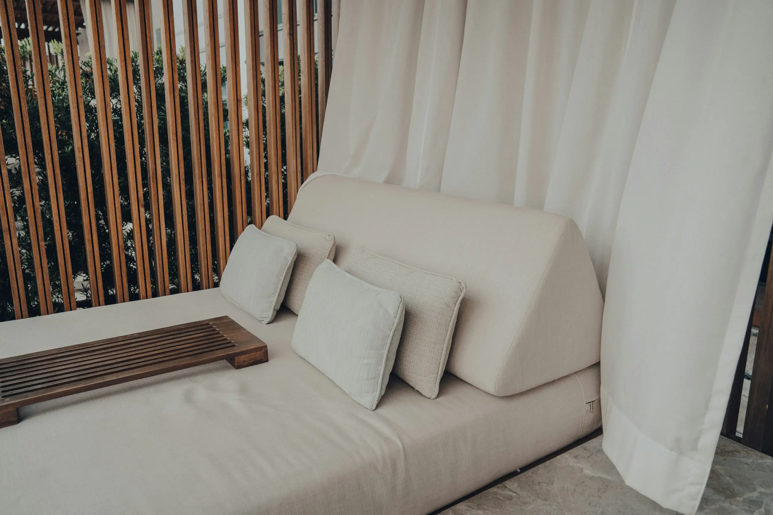 A cream daybed with a bolster cushion, set against a slatted timber screen and linen curtains.
